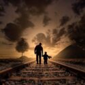 Tumma kuva, vanhempi ja lapsi käsi kädessä kävelevät junaradalla.