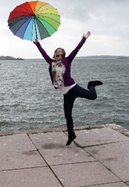 Iloinen, hyppäävä nainen rannalla, kädessään värikäs sateenvarjo.