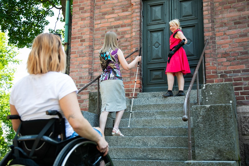 Kolme vammaista henkilöä kirkon portailla, mm. pyörätuoli.
